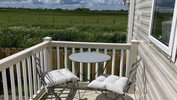 A deck with a table and chairs, a white railing, and a view of a green field. - Remarkable 2-bed Holiday Home in Whitstable (Whitstable)