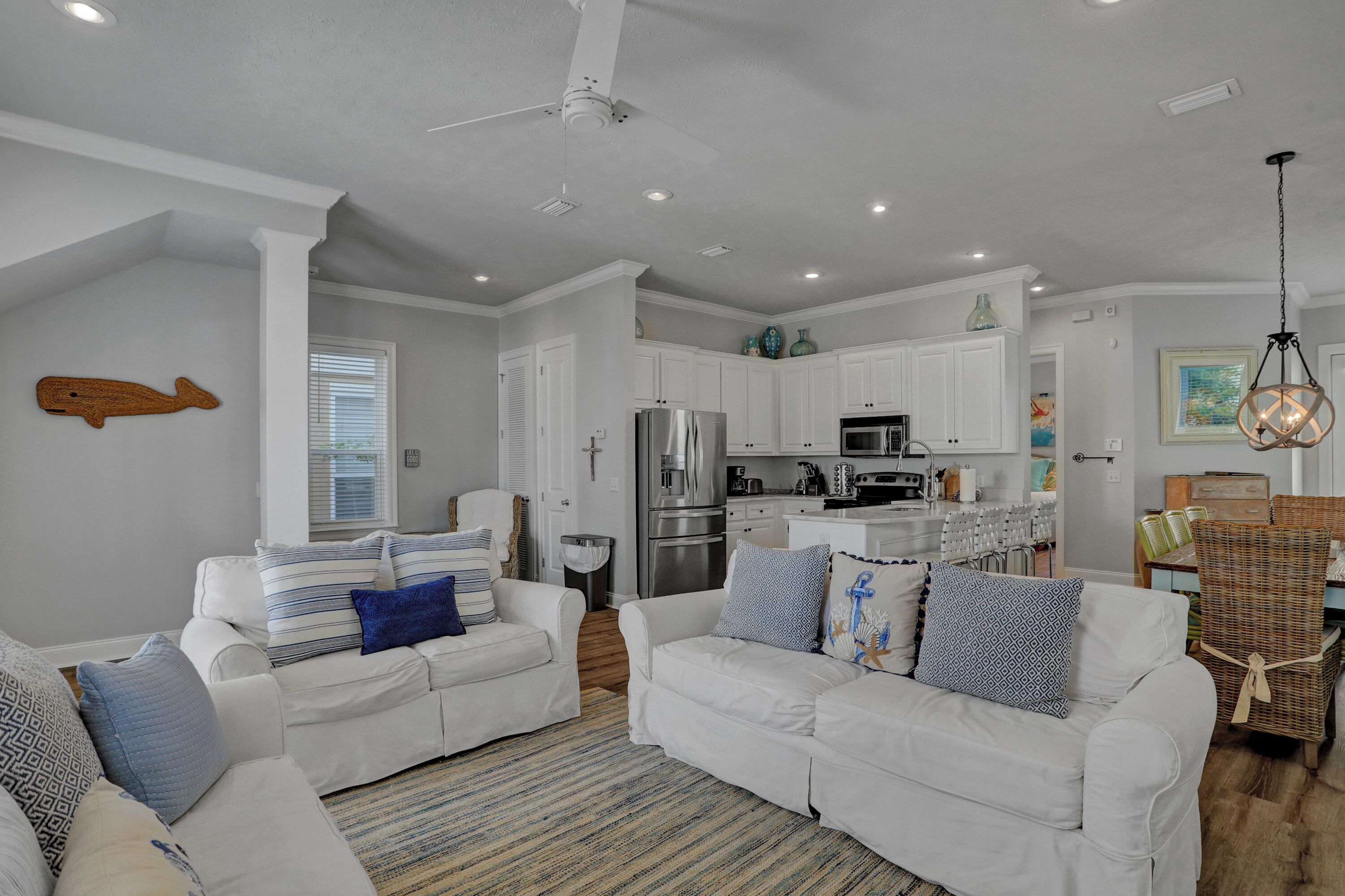 A spacious living room with a white sofa set, a kitchen in the background, and a dining area with wicker furniture.