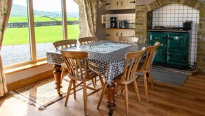 Un comedor con una mesa dispuesta para seis personas, sillas de madera, vistas a un paisaje verde a través de amplias ventanas y una estufa verde en la cocina.