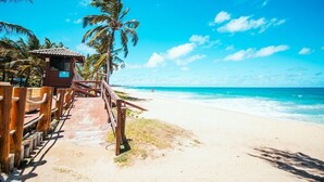 Ein hölzerner Steg führt zu einem Strand mit Palmen und wolkenlosem, blauem Himmel.
