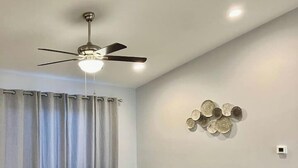 A modern living room with a ceiling fan, light fixture, two beige sofas, a light-colored wooden floor, and a decorative wall piece. - Jake's Cozy Corner (Ocala)