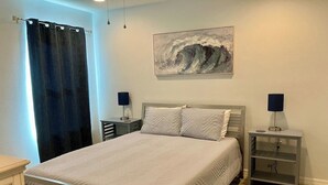A bedroom with a bed, nightstands, a painting, and a window with curtains. - Jake's Cozy Corner (Ocala)