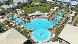 A large, circular swimming pool surrounded by palm trees and lounge chairs, with multiple buildings in the background.