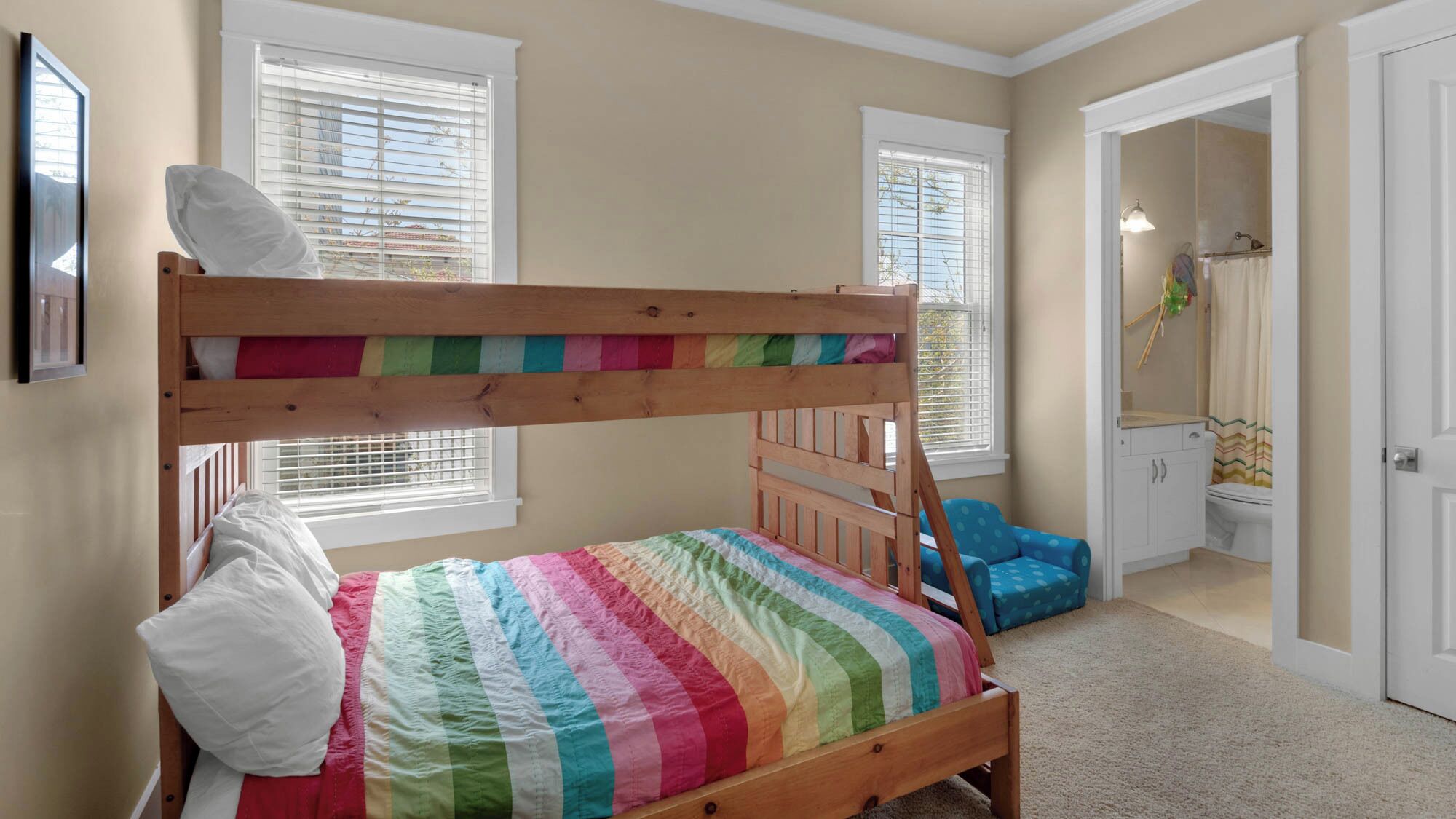 A bunk bed with a colorful striped bedspread, a wooden headboard, and a white pillow. There are two windows with white blinds, a blue chair, and a bathroom visible in the background.