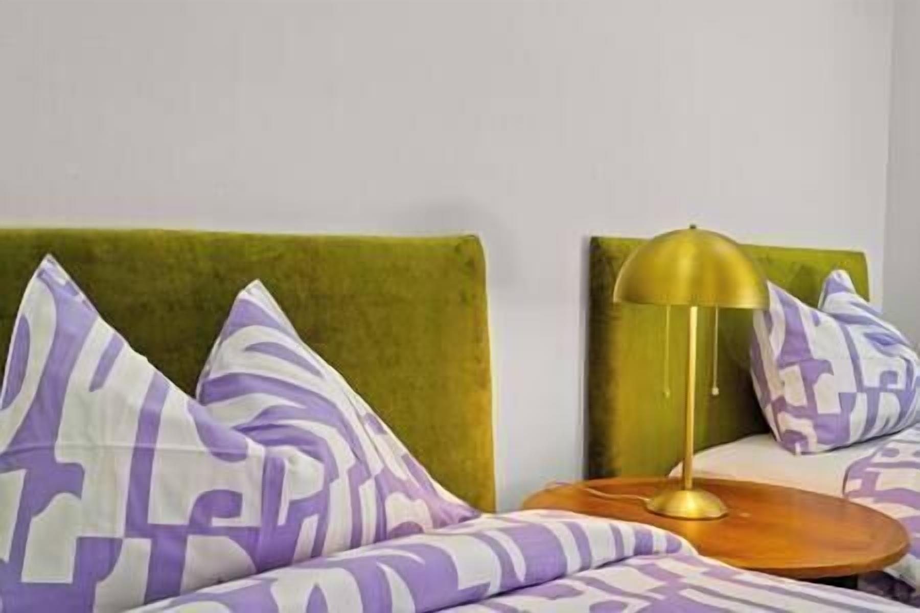 A bed with patterned pillows, a green upholstered headboard, and a bedside table with a lamp.