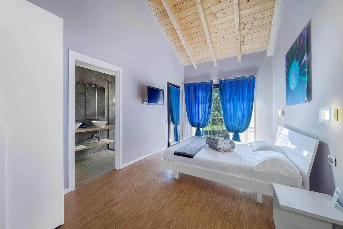 Ein modernes Schlafzimmer mit Holzboden, einem großen Bett, einem Nachttisch, einem Fernseher und einem Badezimmer mit Waschbecken und Spiegel.