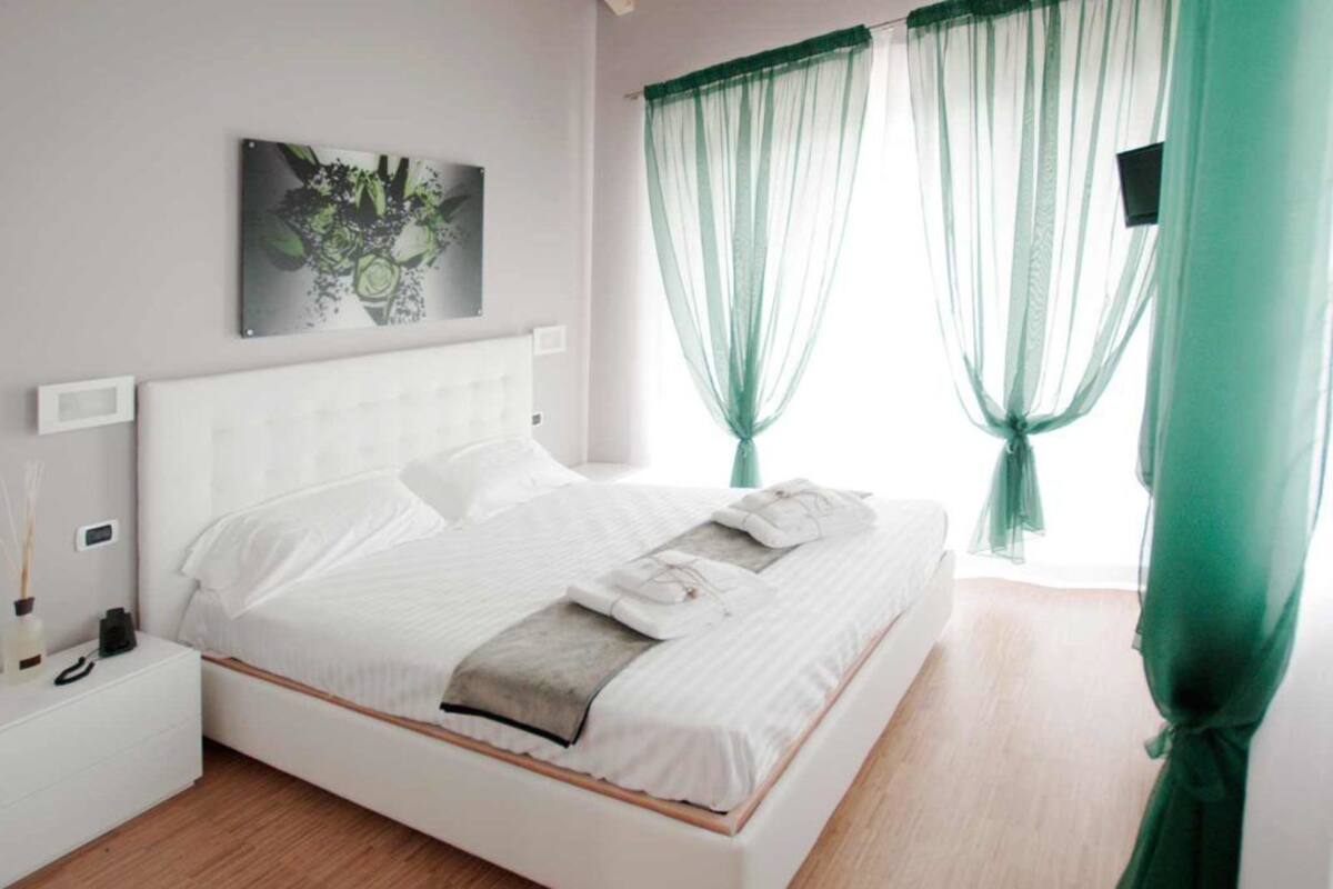 Ein ordentlich bezogenes Bett mit weißen Laken und einer grauen Decke, ein Nachttisch mit Lampe und transparente, grüne Vorhänge.