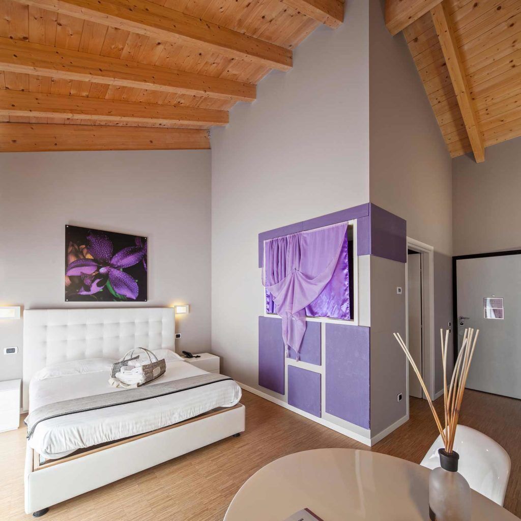 Une chambre à coucher avec un lit, une table de chevet, une étagère fixée au mur et des rideaux violets, ainsi qu’un tableau représentant une fleur.