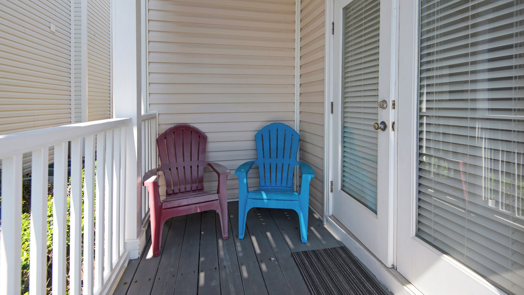 A porch with two Adirondack chairs, a white railing, and a door with blinds.
