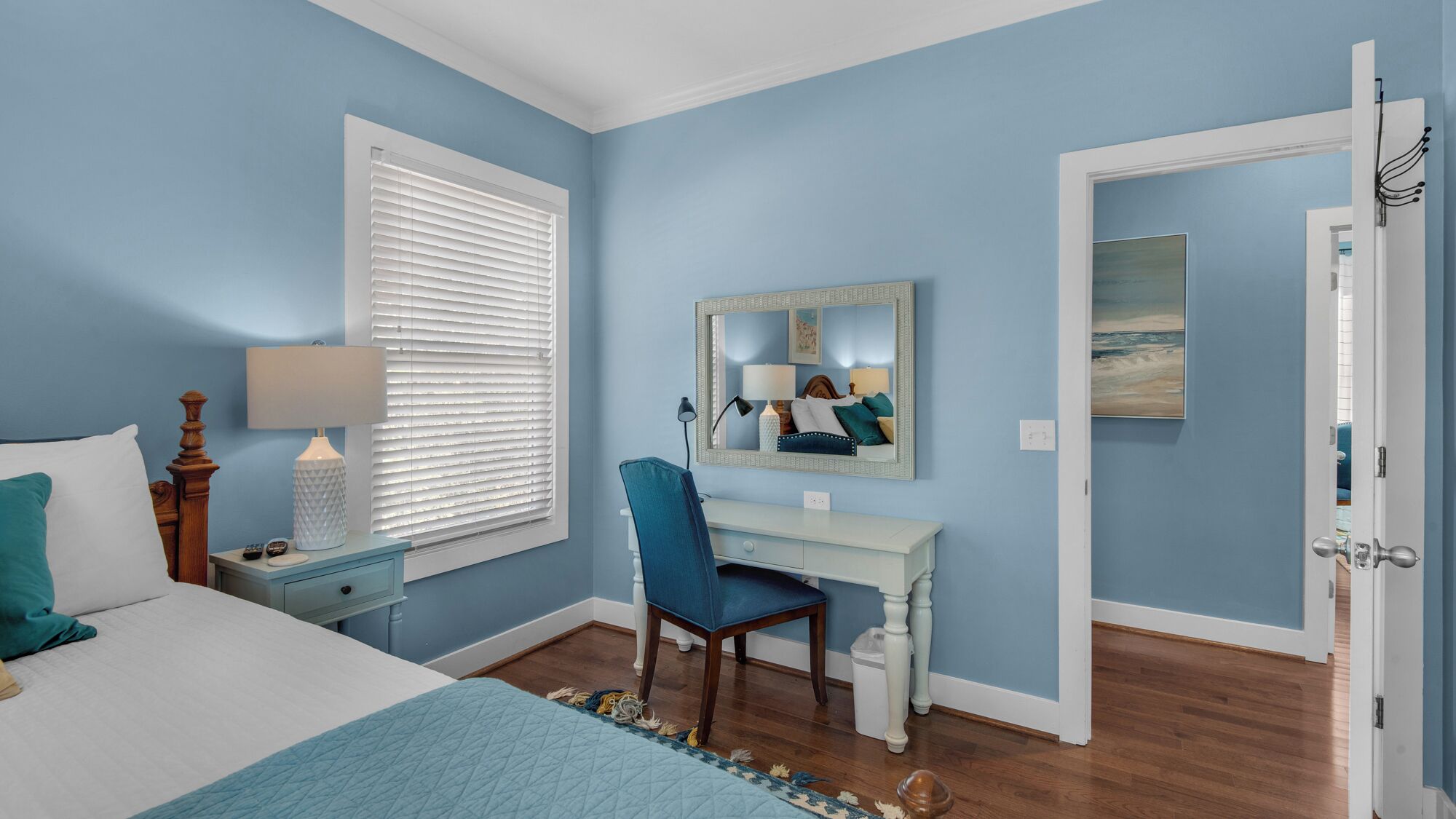 A bedroom with a bed, a desk, a chair, a dresser, a window with blinds, a mirror, and a door.