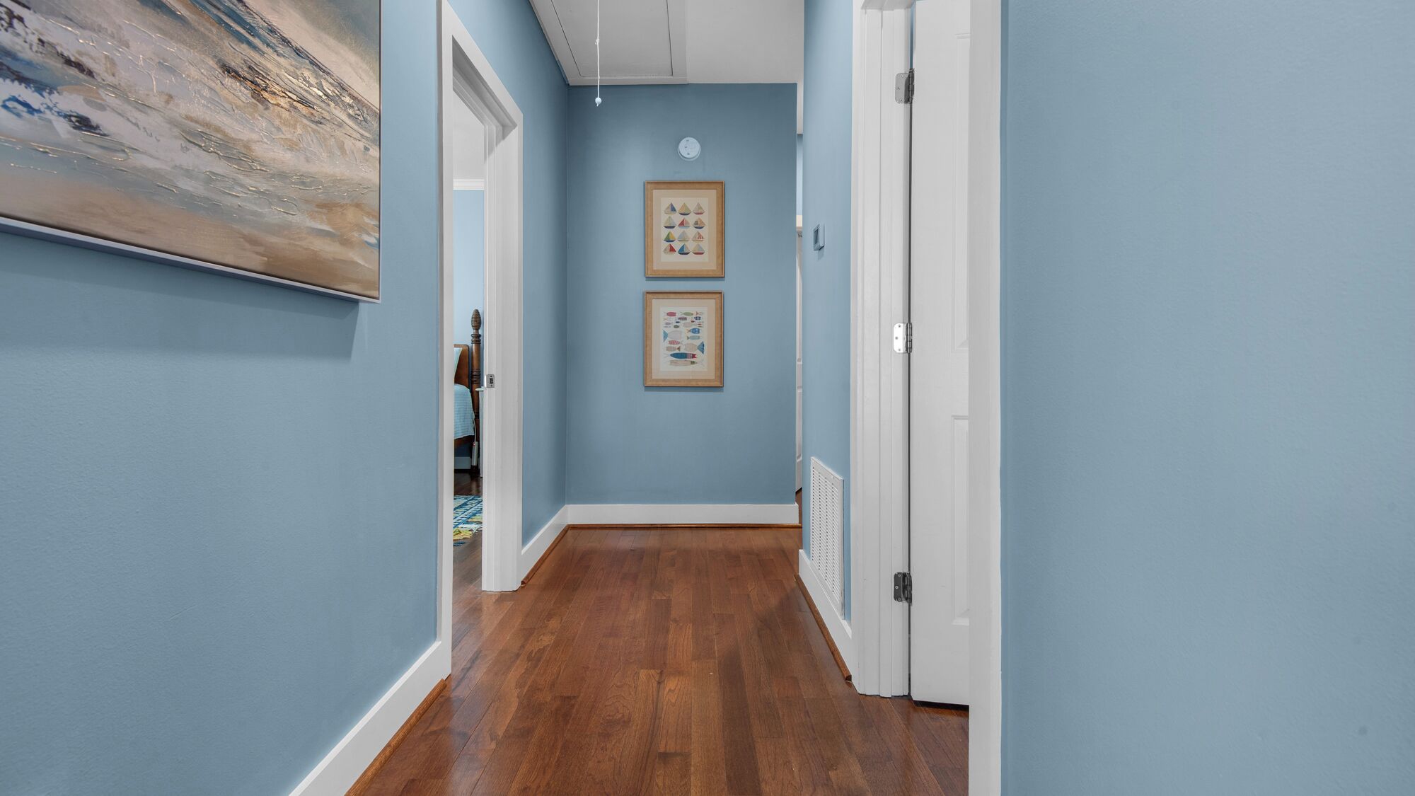 A hallway with blue walls, wooden flooring, and two framed pictures on the wall.