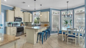 A kitchen with white cabinets, a central island with blue barstools, a dining table with chairs, and a chandelier.