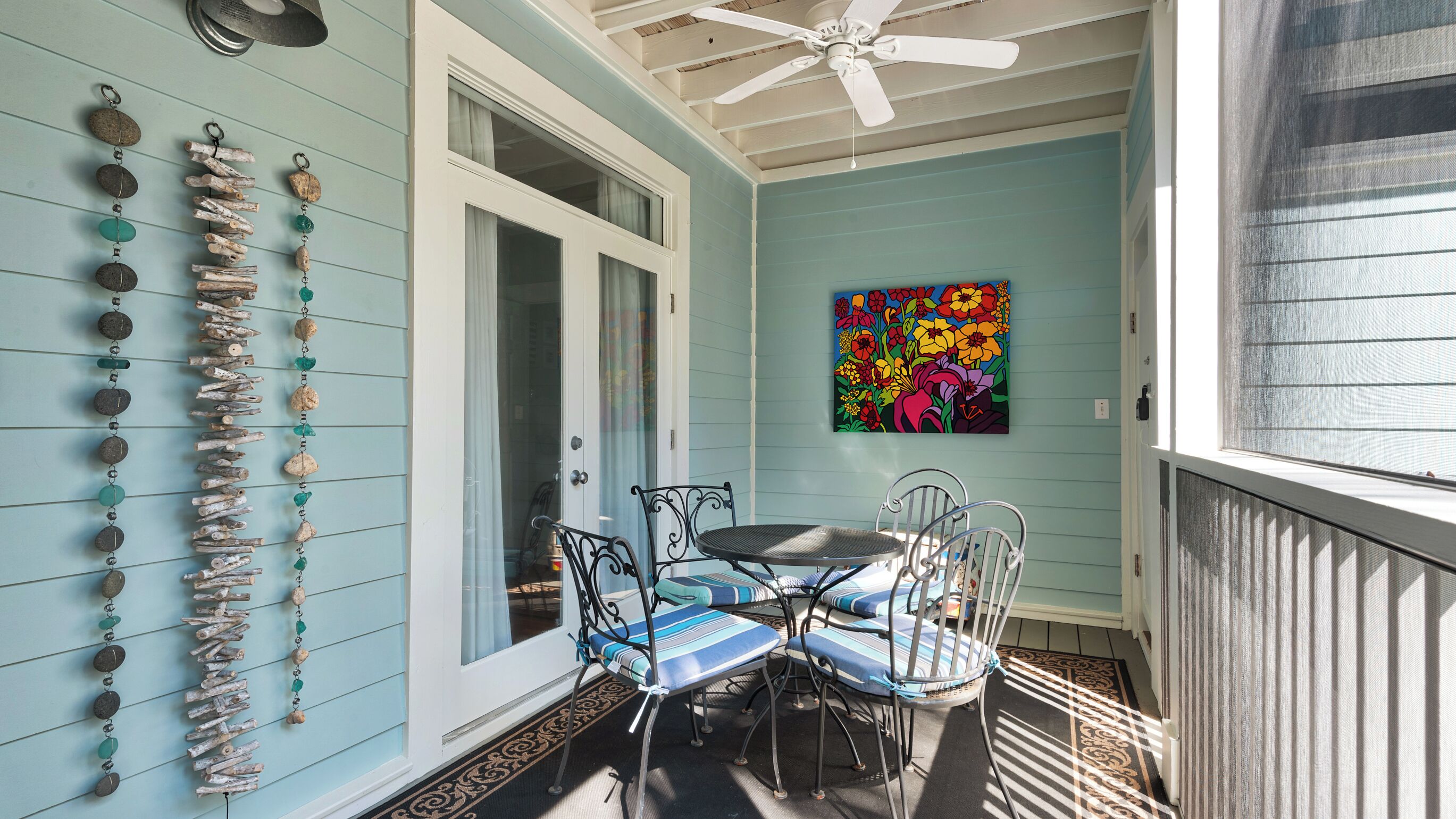 A porch with a ceiling fan, a table with chairs, a colorful painting, and a wall adorned with a string of decorative items.