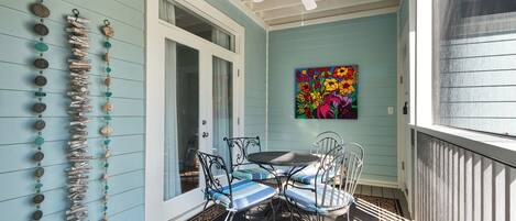 A porch with a ceiling fan, a table with chairs, a colorful painting, and a wall adorned with a string of decorative items.