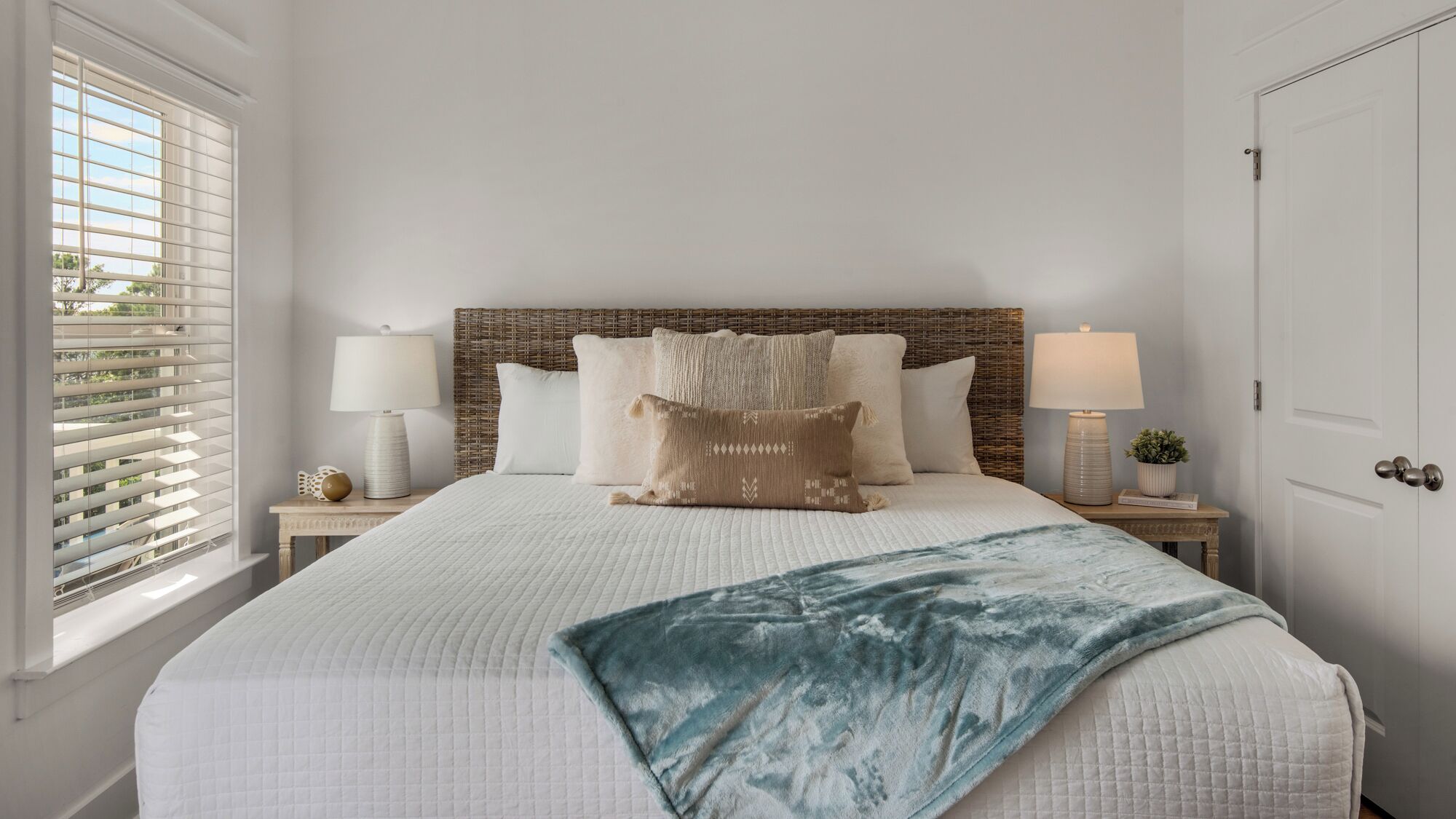 Een net opgemaakt bed met een witte deken, een blauwe plaid en meerdere kussens, waaronder een met een patroon. Twee nachtkastjes met lampen en decoratieve voorwerpen.