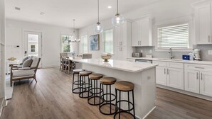 A modern kitchen with a large island, bar stools, and a dining area with a sofa and chairs. - Spacious 4BR With 2 Master Suites, Golf Cart, .3 Miles to Beach - Sleeps 15 (Santa Rosa Beach)