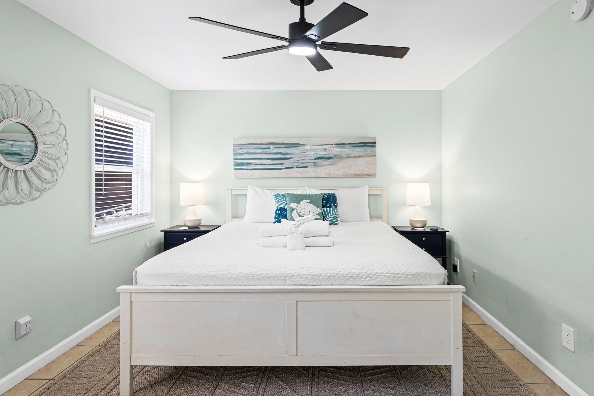 A bedroom with a bed, two bedside tables, a ceiling fan, a window with blinds, and a wall-mounted artwork.