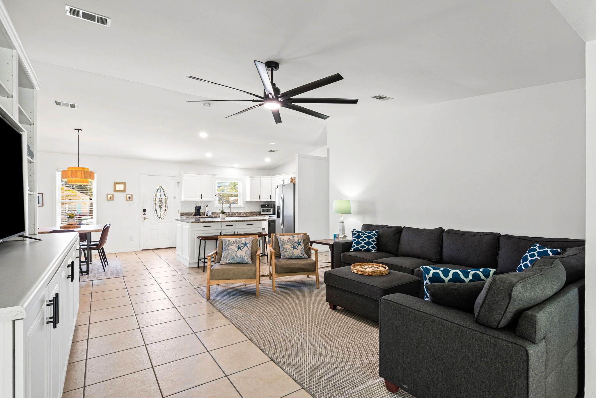 A modern living room with a dark grey sofa, two armchairs, a dining area with a table and chairs, a kitchen in the background, and a ceiling fan.