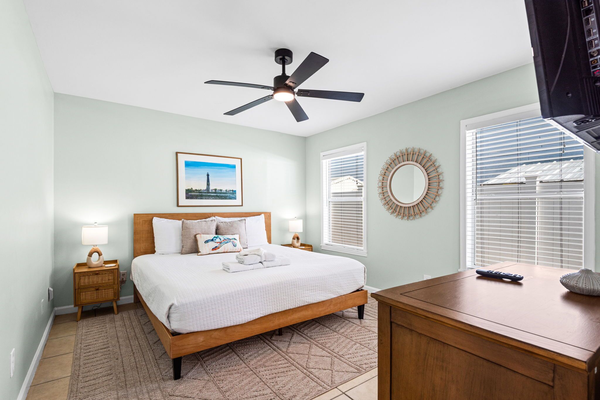 A bedroom with a bed, a ceiling fan, a bedside table with a lamp, a framed picture on the wall, and a window with blinds.