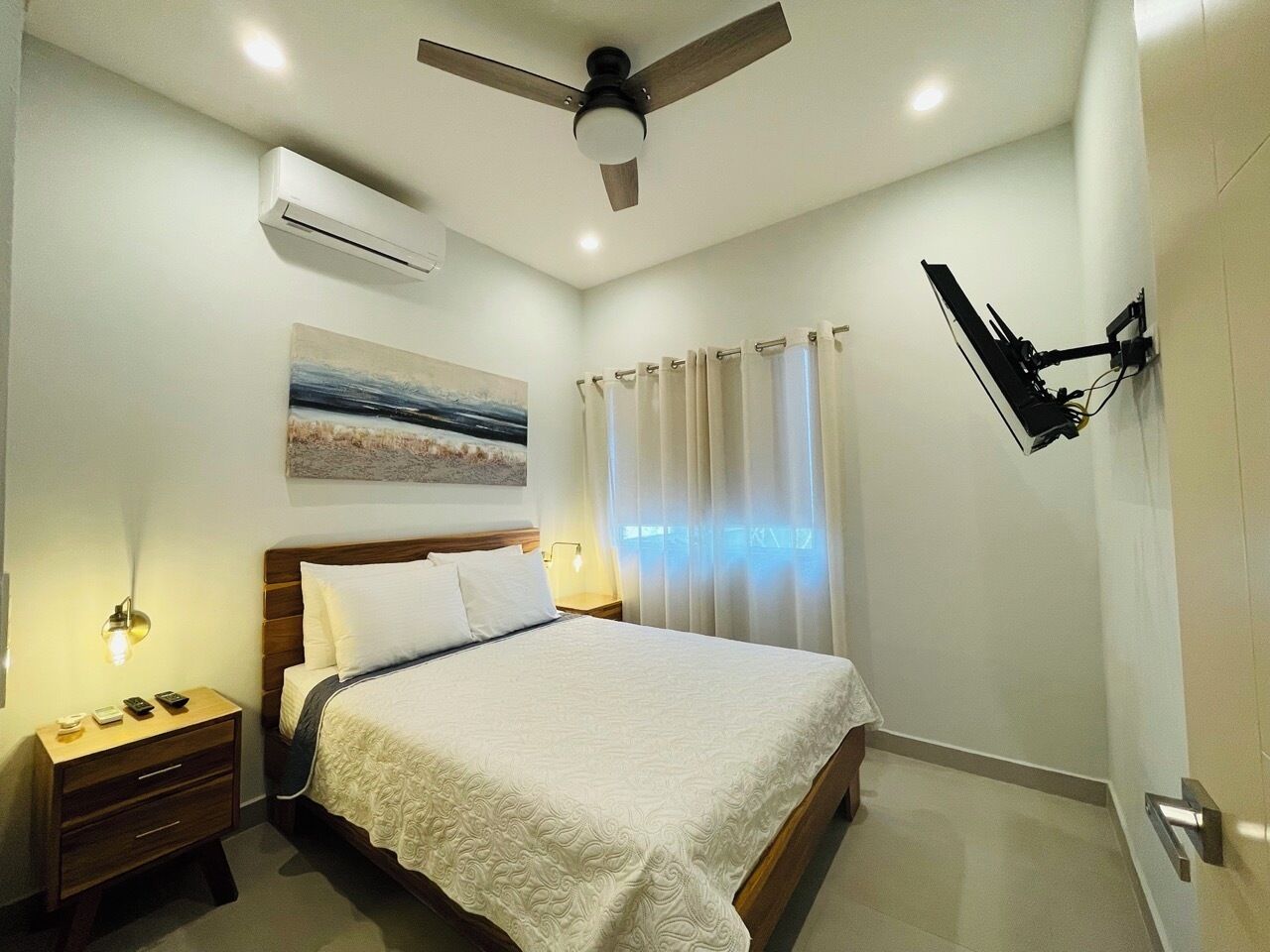 A bedroom with a bed, nightstand, wall-mounted TV, ceiling fan, and an air conditioning unit.