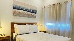 A bedroom with a wooden bed, white bedding, a nightstand with a lamp, a wall-mounted air conditioner, and a beach-themed painting. - Casa Lapa Roja (Jacó)