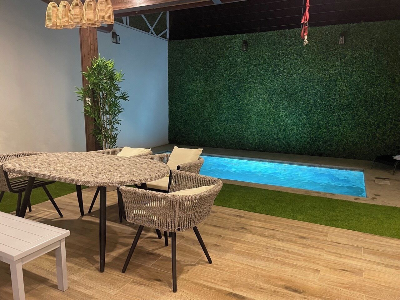 Uma área à beira da piscina com mesa e cadeiras de vime, uma pequena mesa branca e um painel de grama sintética verde.
