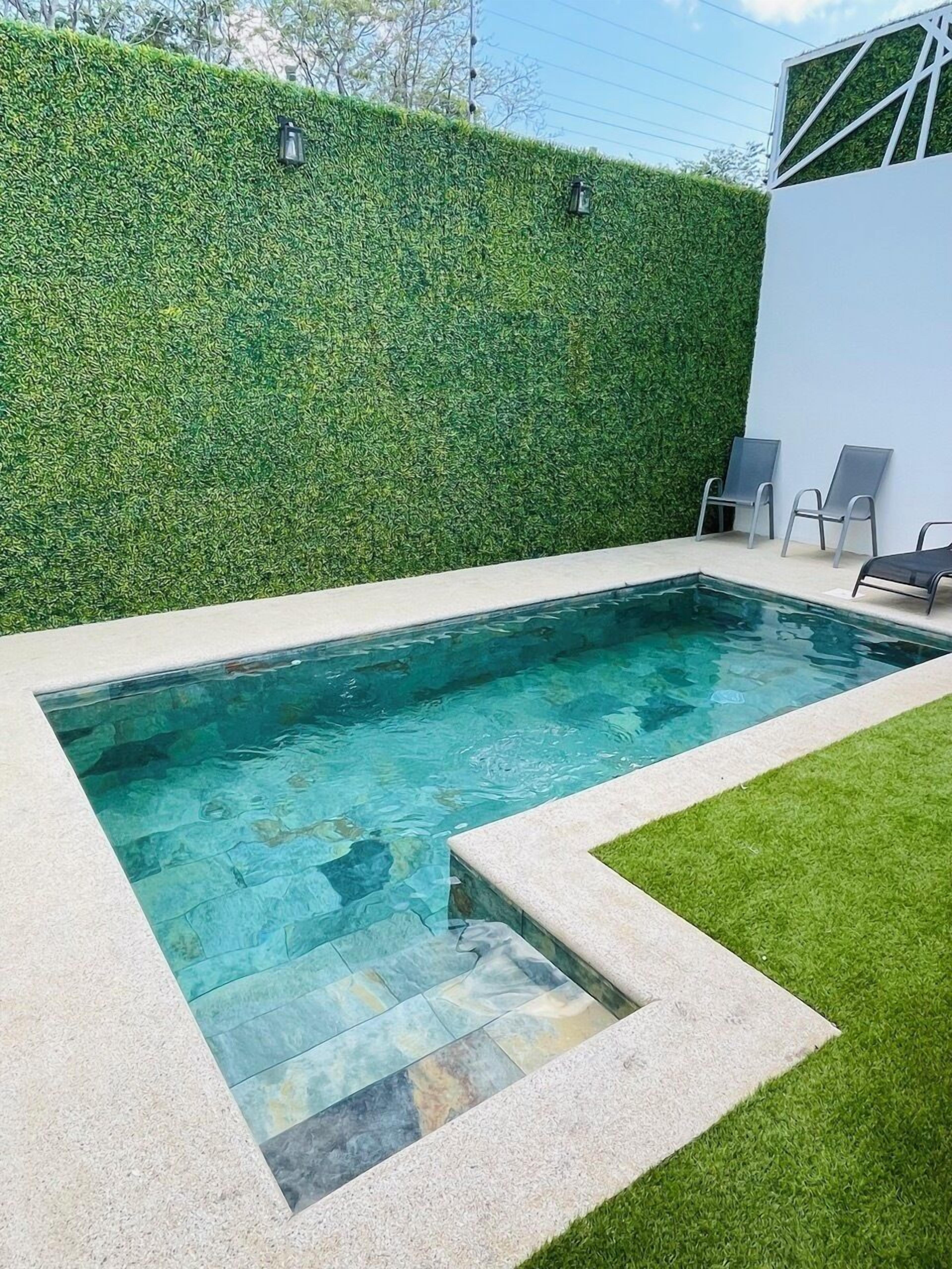 A rectangular swimming pool with clear blue water, surrounded by a low stone border. Adjacent to the pool is a well-maintained grassy area with a hedge wall and two metal chairs.