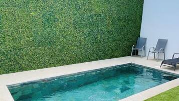 A rectangular swimming pool with clear blue water, surrounded by a low stone border. Adjacent to the pool is a well-maintained grassy area with a hedge wall and two metal chairs.