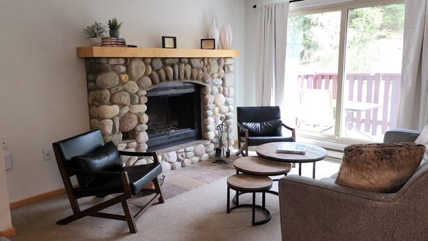 A cozy living room with a stone fireplace, leather chairs, a round coffee table, and a view of a wooden deck through a large window. - 3 Bedroom Centrally Located Ski In/Ski Out Condo in Panorama (Panorama)