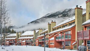 A row of red wooden buildings with snow-covered roofs, set against a backdrop of a mountain and mist. - 3 Bedroom Centrally Located Ski In/Ski Out Condo in Panorama (Panorama)
