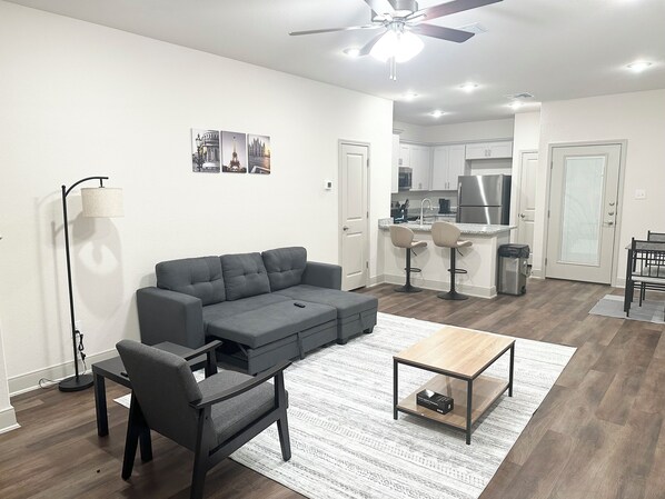 A modern living room with a grey sectional sofa, a coffee table, and a dining area with two bar stools. A kitchen with white cabinets and stainless steel appliances is visible in the background.