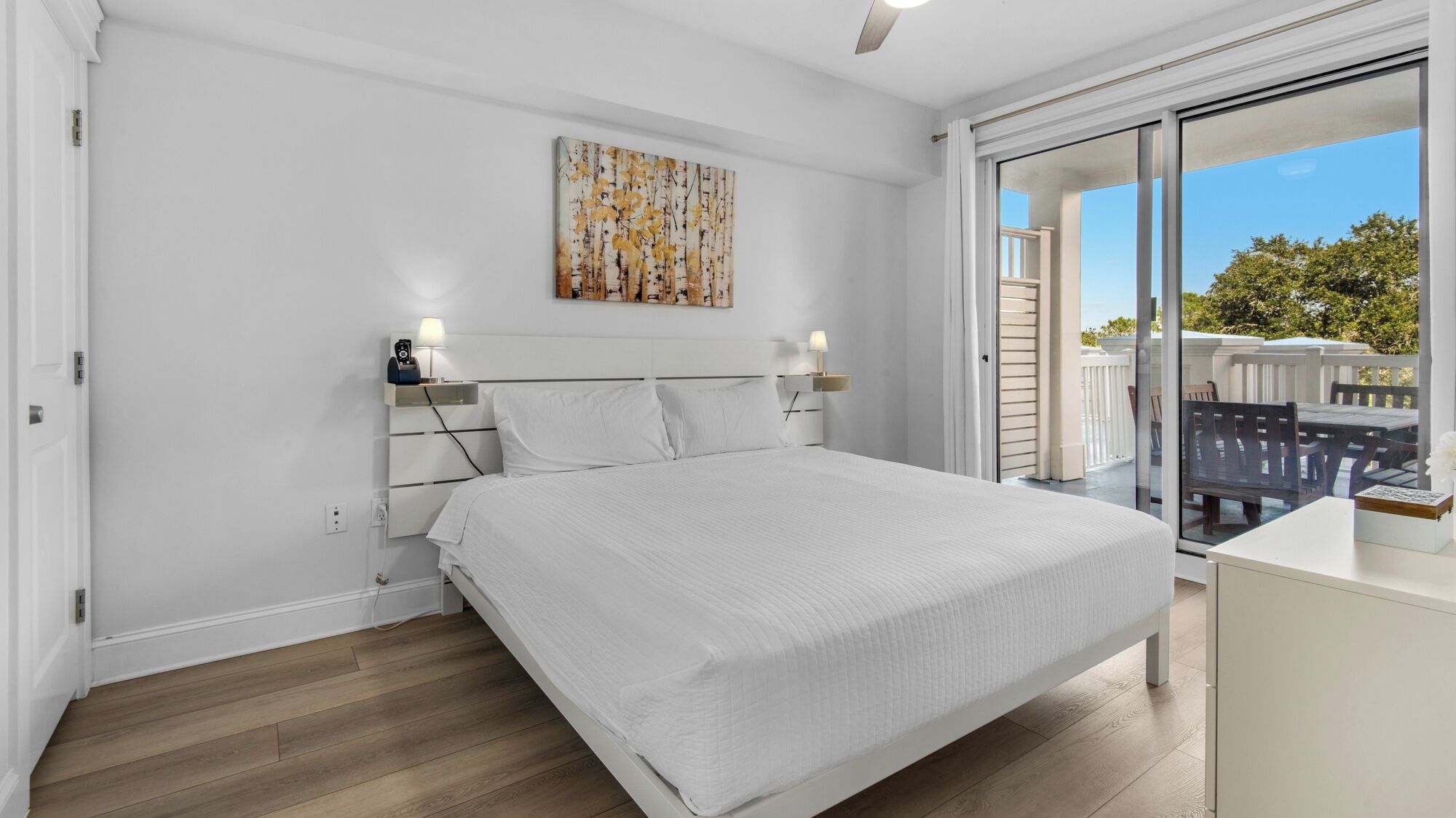 Un dormitorio con una cama grande, una mesita de noche con lámpara, un cuadro en la pared y vistas al exterior a través de un ventanal corredero.