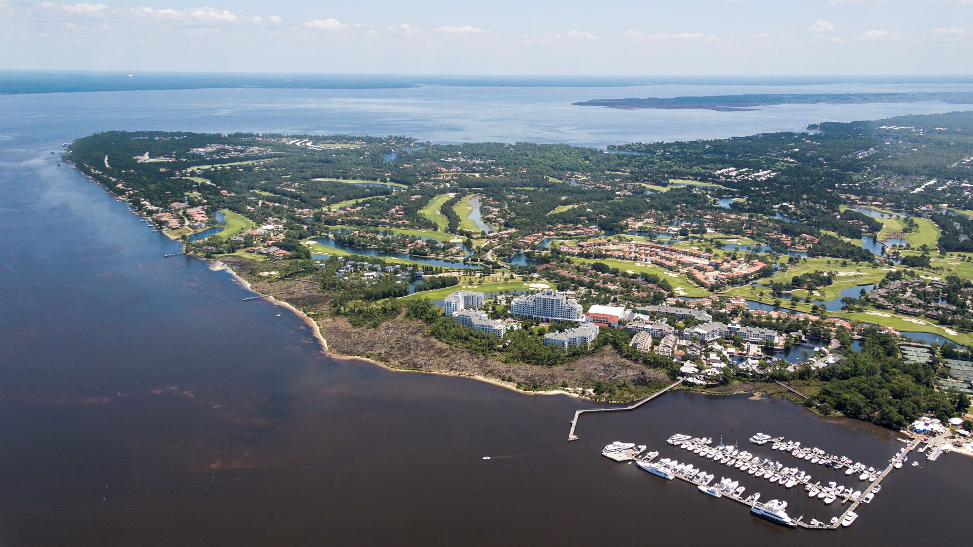 An aerial view of a coastal area with a marina, residential buildings, and golf courses.