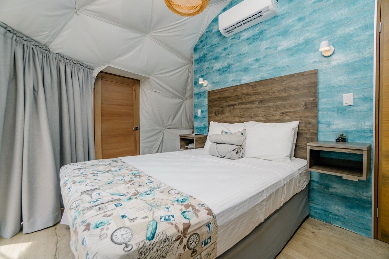A bedroom with a bed, a wooden headboard, a nightstand, a wall-mounted shelf, and a window with curtains.