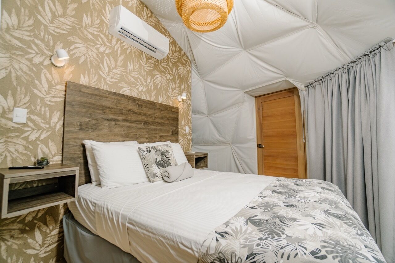 A bedroom with a wooden headboard, a bed with white bedding, a wall-mounted air conditioner, a bedside lamp, and a hanging light fixture.