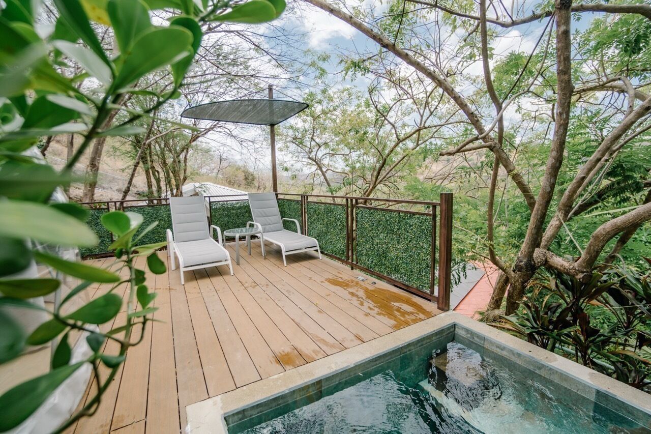 Terraza de madera con dos sillones blancos y una mesa, rodeada de follaje y una piscina.