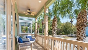 A covered porch with a ceiling fan, wicker furniture, and a view of palm trees and a parking lot.