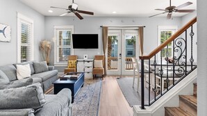 A living room with a sofa, coffee table, and a TV. There are two ceiling fans, a staircase with a railing, and a door leading to a balcony.