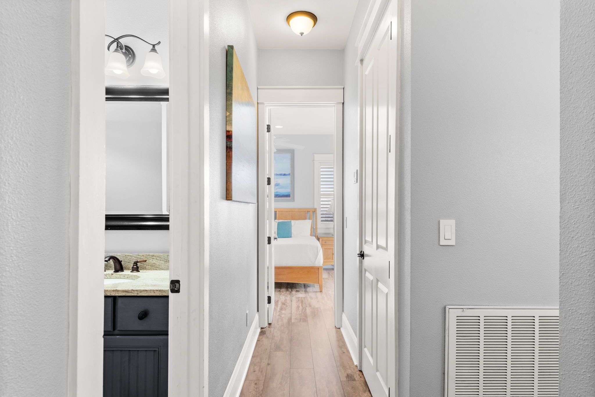 A narrow hallway with a bathroom on the left, a bedroom with a bed and window in the center, and a door leading to another room on the right.