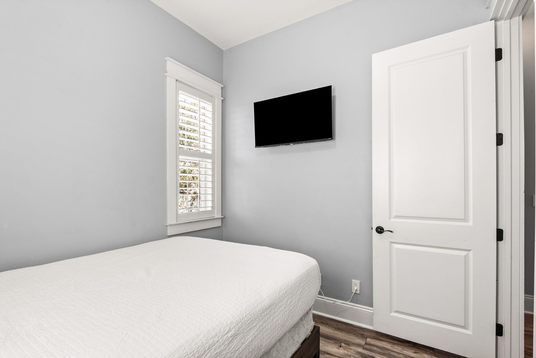 A bedroom with a bed, a window with blinds, a television on the wall, and a door.