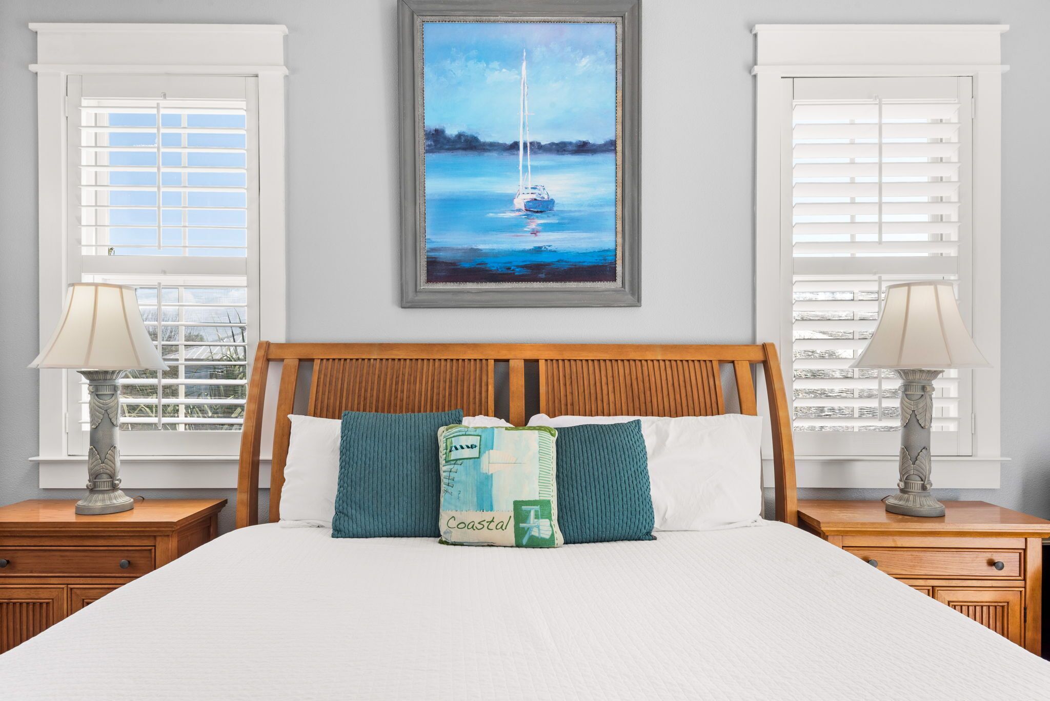 A bedroom with a bed, two nightstands, a painting of a sailboat, and a window with blinds.