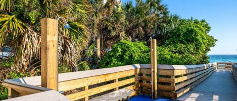 A wooden boardwalk with benches, surrounded by lush greenery and palm trees, leading to a beach.