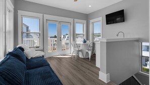 A modern living room with a blue sofa, a dining area with a white table and chairs, a kitchen with a sink and a TV mounted on the wall. - Steps From the Seagrove Beach - Gulf Views - Sleeps 10 - Community Pool (Santa Rosa Beach)