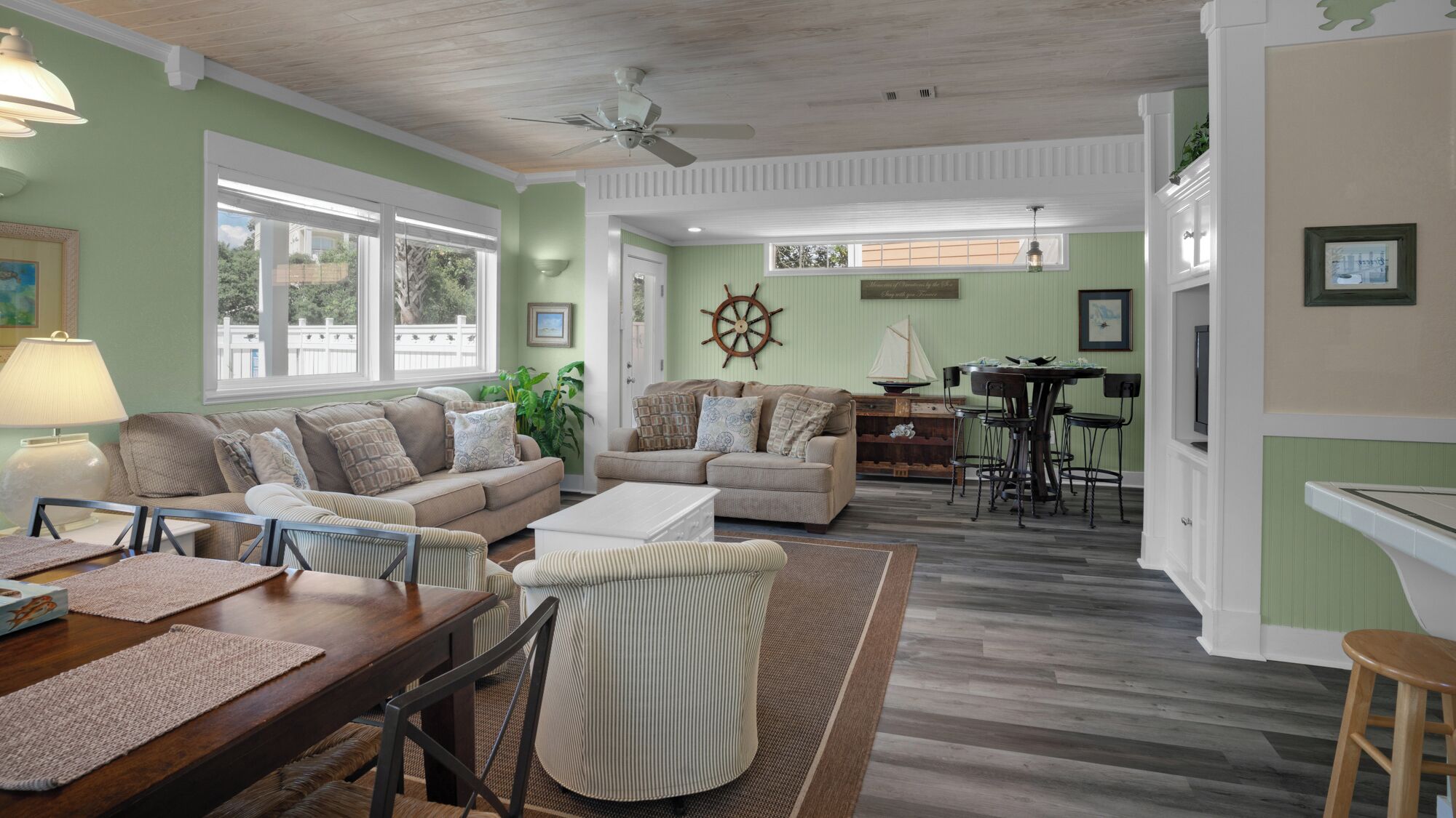 A spacious living room with a dining area, featuring a large window, a ceiling fan, and a sailboat decoration.
