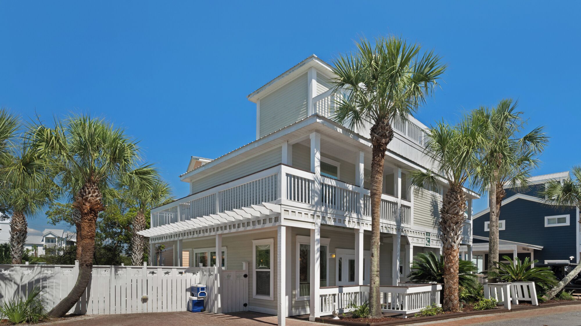 A two-story beachfront house with a balcony, surrounded by palm trees.