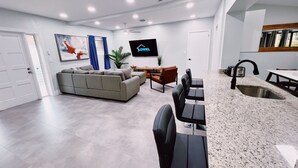 A modern living room with a grey sofa, a dining area with a granite countertop and bar stools, and a flat-screen TV mounted on the wall.
