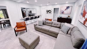 A modern living room with a grey sofa, a brown leather chair, and a glass coffee table. There is a kitchen area with a bar and seating.