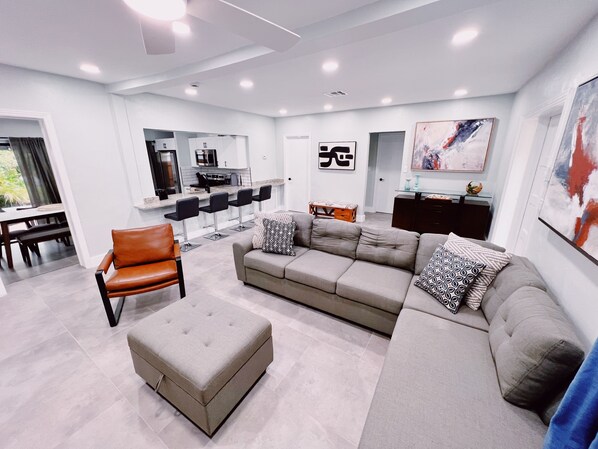 Ein modernes Wohnzimmer mit einem grauen Sofa, einem braunen Ledersessel und einem Glastisch. Es gibt eine Küchenzeile mit einer Bar und Sitzgelegenheiten.