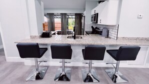 A modern kitchen with a granite countertop, four black bar stools, a built-in microwave, and a view of the backyard through a glass door.