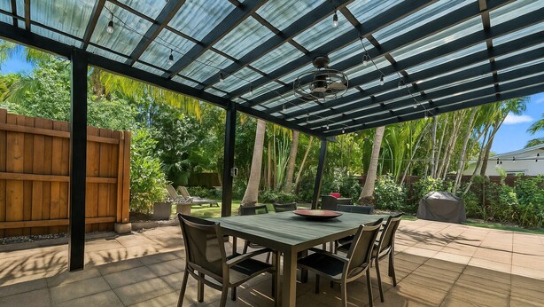 A covered patio with a table and chairs, surrounded by lush greenery and a wooden fence.